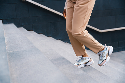 A woman in a beige trousers and sneakers ascends the stairs, we see only her legs in a daylight