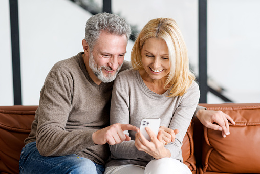Happy senior couple with smartphone on the couch spends leisure time in social networks, scrolling feed, watching videos, grey-haired man is showing smth to smiling beautiful woman