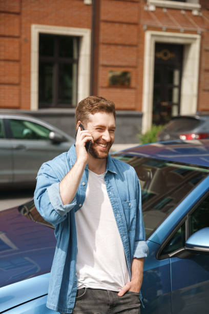istock A young handsome driver in a jeans outfit talks on his phone with a smile, leaning on a blue car in the sunlight 1352269683