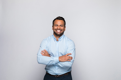Portrait of handsome mid adult man wearing blue shirt, standing with arms crossed and smiling at camera. Studio shot of male entrepreneur against grey background.