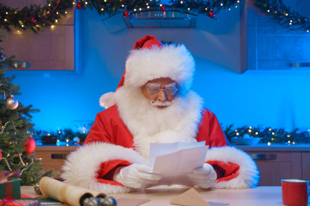 istock Serious Santa Claus reads a letter, sitting at a table with a scroll and envelopes on it in a decorated kitchen with Christmas lights 1352271204