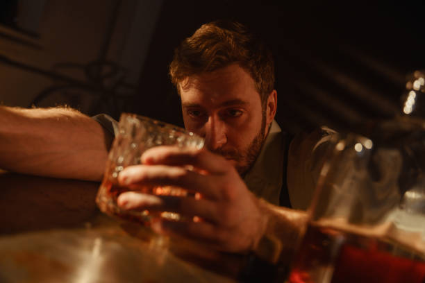 atmospheric portrait of a drunk man with a glass of whiskey at the table. a man in a strong alcoholic intoxication looks at a glass of whiskey, which he holds in his hand. - intoxication imagens e fotografias de stock