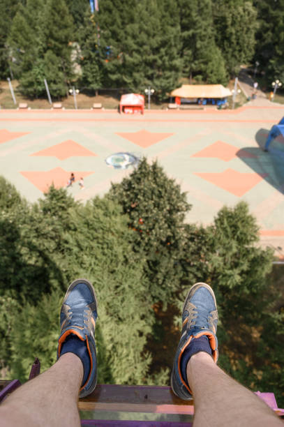 From above anonymous man in sneakers riding modern Ferris wheel over green trees on fairground in summer