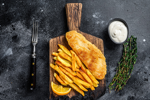 Fish and chips dish with french fries on wooden board. Black background. Top view.