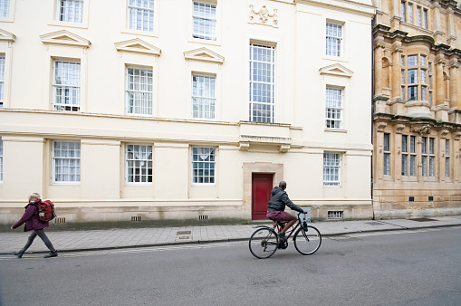 A cyclist and a senior man walking along Broad Street in Oxford, Oxfordshire, England, UK in front of Georgian buildings.