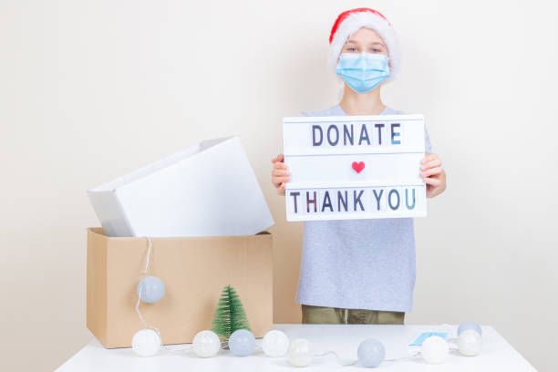 christmas donation, charity. kid volunteer with santa hat and medical protective face mask holding lightbox with message donate and thank you. boy standing near table with empty cardboxes for donation - charity and relief work donation box thank you child imagens e fotografias de stock