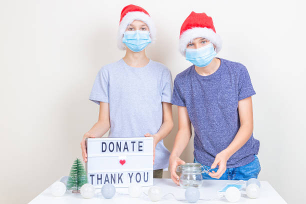 teenager boys volunteers with protective face mask preparing to collect christmas donations indoors. kids standing near desk with christmas decorations and lightbox with message donate and thank you - charity and relief work donation box thank you child imagens e fotografias de stock