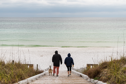 Seaside, USA - January 10, 2021: Couple people with dog walking in Seaside, Florida gulf coast beach with wooden boardwalk steps down and white sand with colorful emerald water