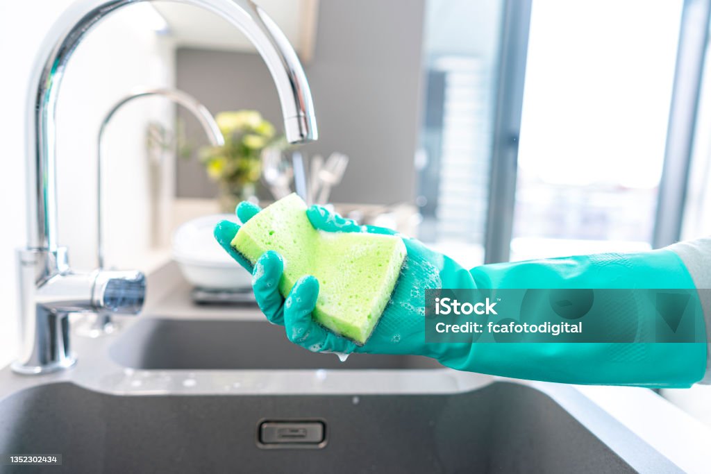 Woman hand with glove holding wet cleaning sponge Close up of woman hand wearing a green washing up glove holding a wet cleaning sponge on a kitchen sink. A faucet is visible. High resolution 42Mp indoors digital capture taken with SONY A7rII and Zeiss Batis 25mm F2.0 lens Cleaning Sponge Stock Photo