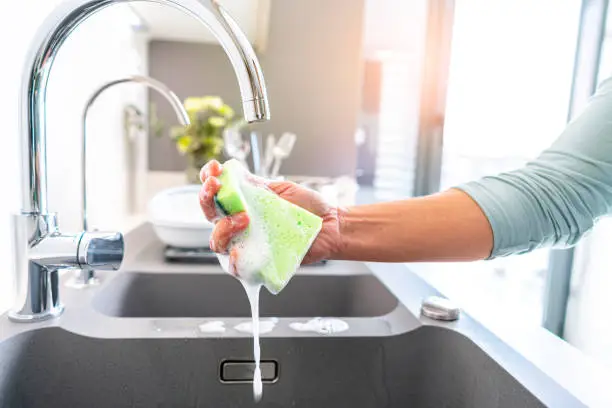 Close up of woman hand holding a wet cleaning sponge on a kitchen sink. A faucet is visible. High resolution 42Mp indoors digital capture taken with SONY A7rII and Zeiss Batis 25mm F2.0 lens