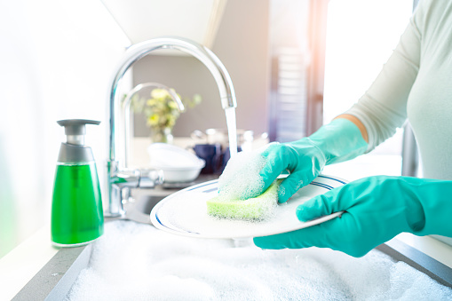 Close up of woman hands  washing a plate with a cleaning sponge in a kitchen sink. A faucet with flowing water and a dish washing soap are visible. The sink is full of water and soap sud. High resolution 42Mp indoors digital capture taken with SONY A7rII and Zeiss Batis 25mm F2.0 lens