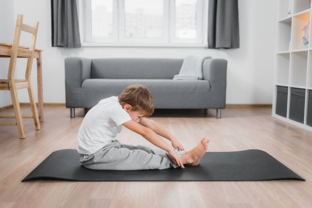 istock Boy child makes a forward bend from a sitting position on the floor with straight legs, wearing a white t-shirt and gray pants. 1352303416