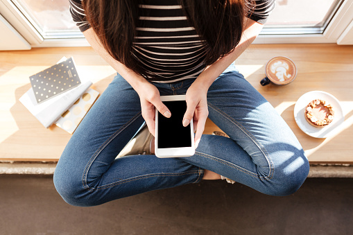Top view of Asian woman which sitting on windowsill with phone and cup of coffee. Cropped image