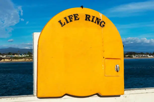 Life Ring cabinet with yellow cover on pier railing. Blurred scenic ocean coast background.