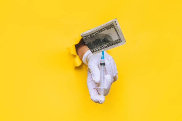 A doctor's hand in a white medical glove holds a syringe with a vaccine on which a dollar bill is pinned. The concept of financial costs and income from vaccination. Fighting coronavirus, covid-19. stock photo