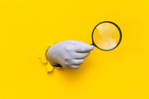 Doctor's hand in a white latex medical glove holds a magnifying glass on a yellow background. Torn paper with copy space. Selective focus. Search, zoom and research concept. stock photo