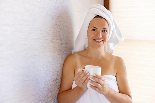 Attractive young girl in bath towel and with a towel on her head holds a cup and smiles.