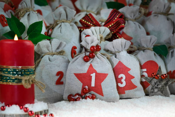 close-up advent calendar diy sacks of gray canvas tied with twine,blurred candle on side,christmas decor,red berries,wooden star,poinsettia leaves and snow in foreground. celebrating and gifts concept - burlap canvas home decorating color image imagens e fotografias de stock