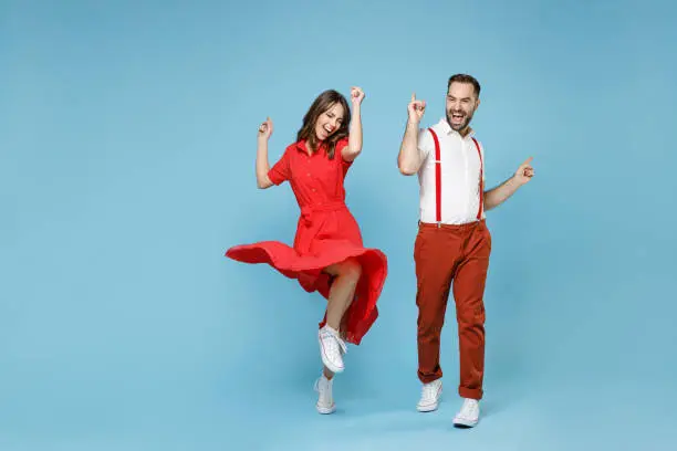 Photo of Full length of cheerful young couple friends man woman in white red clothes dancing point fingers up having fun isolated on pastel blue background studio portrait. St. Valentine's Day holiday concept.