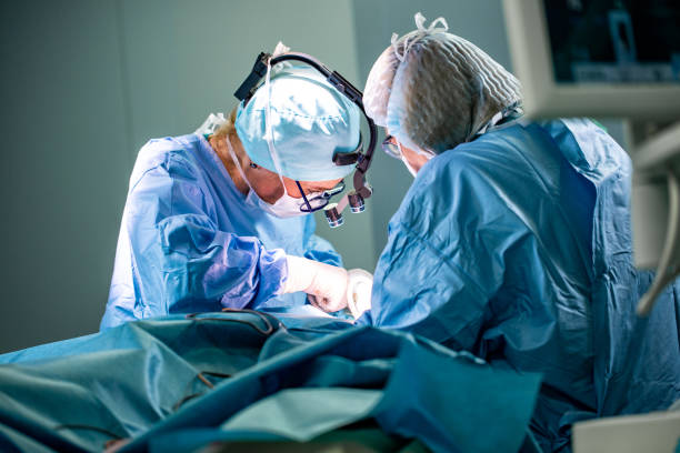 surgeon and his assistant performing cosmetic surgery in hospital operating room. surgeon in mask wearing loupes during medical procadure - cirurgia plástica imagens e fotografias de stock