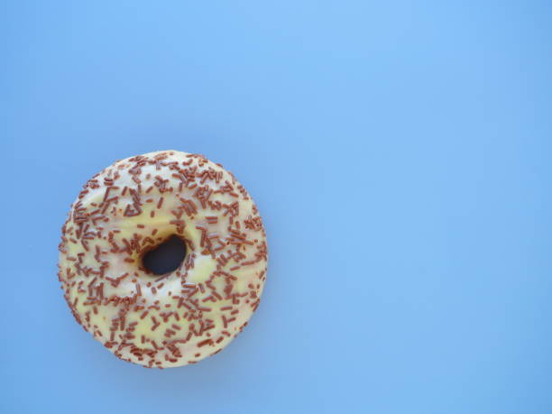 istock Doughnut close-up on blue background. Delicious cake covered with vanilla icing and chocolate sprinkles. High-calorie breakfast or snack. Sweet dessert with vanilla and chocolate, donut or dough. 1352345383