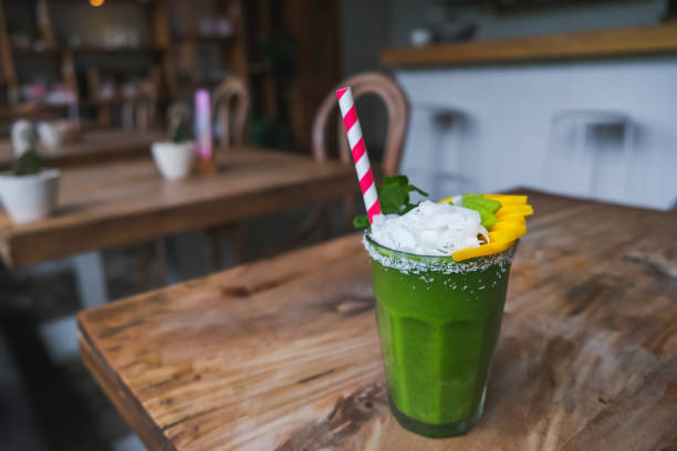 Close-up shot of healthy green smoothies with slice of mango fruit, kiwi fruit and coconut flakes on top. There is a striped paper straw inside drinking glass.