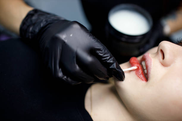 istock Cosmetologist applying red permanent make up tattoo on young woman lips. Permanent lips tattoo procedure concept. 1352346752