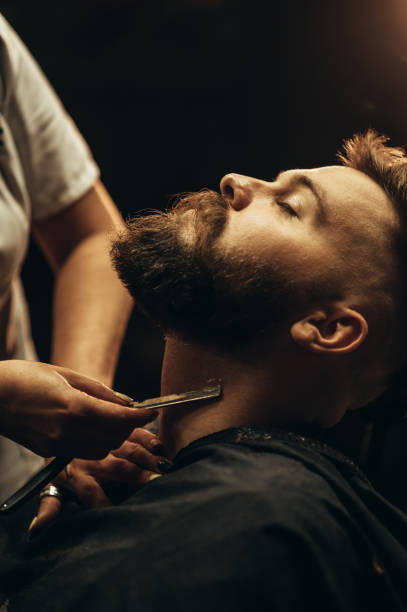 Man getting shaved with straight edge razor by hairdresser at barbershop Young bearded man getting shaved with straight edge razor by hairdresser at barbershop barber shop stock pictures, royalty-free photos & images