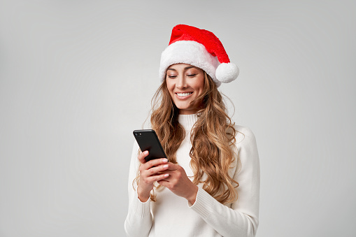 Woman christmas Sants Hat white sweater white studio background with smartphone in hand Beautiful caucasian female curly hair portrait. Happy person positive emotion Holiday concept Teeth smiling