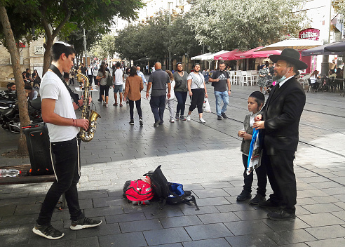 A young street musician playing his saxophone in the center of Jerusalem while a rabbi and his son are watching the show.