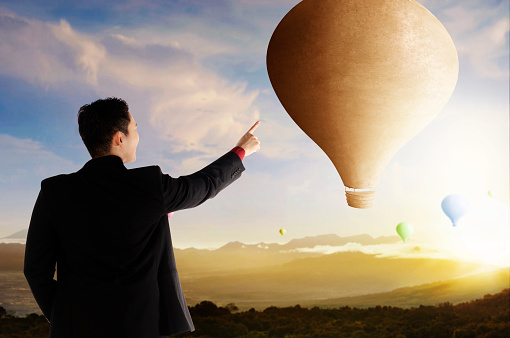Rear view of Asian businessman pointing colorful air balloon flying with sunset sky background