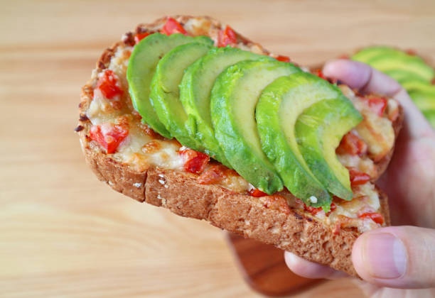 Closeup a delectable grilled cheese toast topped with tomato and sliced avocado in hand
