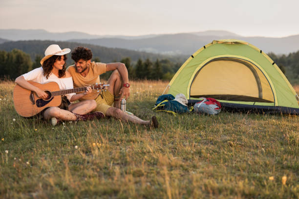 Happy woman playing a guitar for her boyfriend during camping in nature. Copy space.