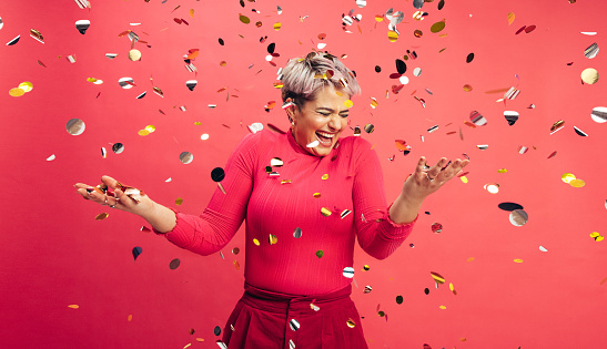 Time to celebrate. Carefree young woman getting excited about winning while standing under falling confetti in a studio. Vibrant young woman celebrating life and having fun against a red background.