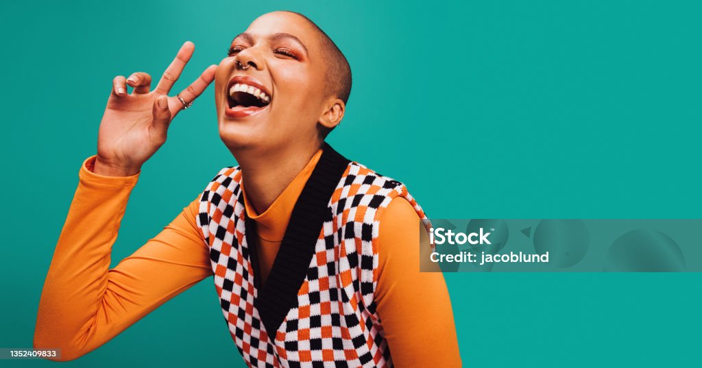 Carefree young woman showing the peace sign in a studio Carefree young woman showing the peace sign in a studio. Happy young woman laughing with her eyes closed while standing against a turquoise background. Woman with septum ring feeling cheerful. Generation Z Stock Photo