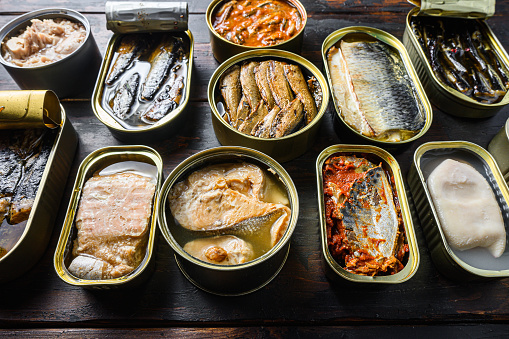 Opened preserve cans with  Saury, mackerel, sprats, sardines, pilchard, squid, tuna over vintage homemade wood table close up side view.