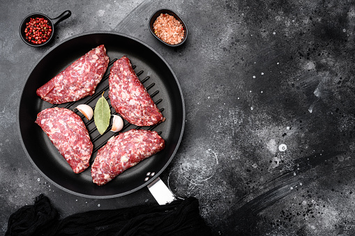 Organic raw ground beef, round patties set, in cast iron frying pan, on black dark stone table background, top view flat lay, with copy space for text