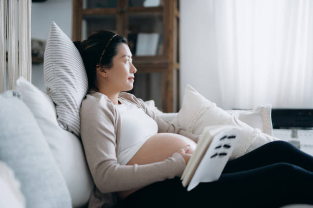 азиатская беременная женщина отдыхает в уютном доме, задумчиво смотрит во время чтения детской книжки на диване. ожидание новой жизни, роды - new childbirth new life love стоковые фото и изображения