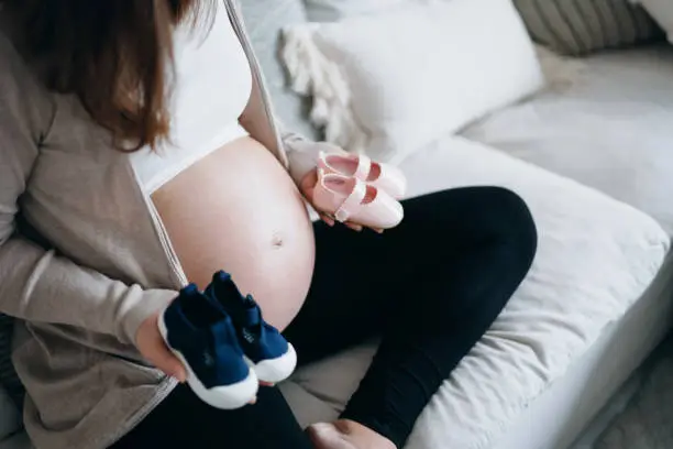 Cropped shot of Asian pregnant woman holding a pair of blue and pink baby shoes in front of her belly. Expecting a new life, mother-to-be, gender reveal concept