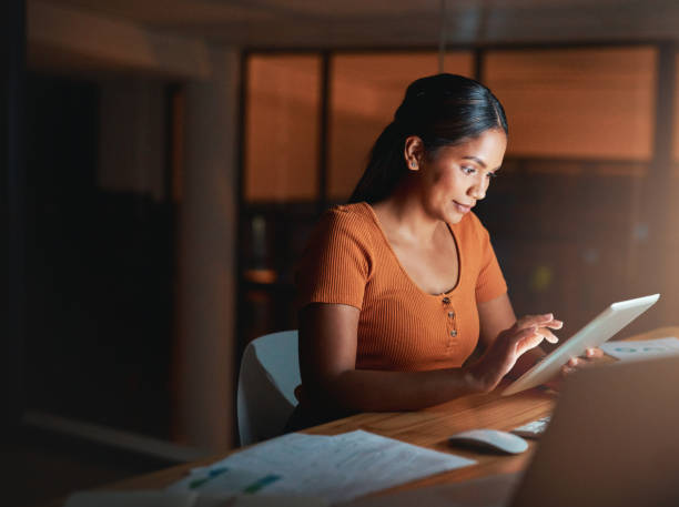 Shot of an attractive young businesswoman sitting alone in the office at night and using a digital tablet Everything seems to be going according to plan indian ethnicity stock pictures, royalty-free photos & images