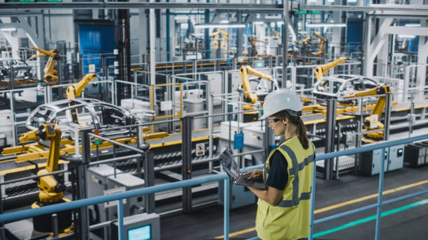 istock Female Car Factory Engineer in High Visibility Vest Using Laptop Computer. Automotive Industrial Manufacturing Facility Working on Vehicle Production with Robotic Technology. Automated Assembly Plant. 1352428669