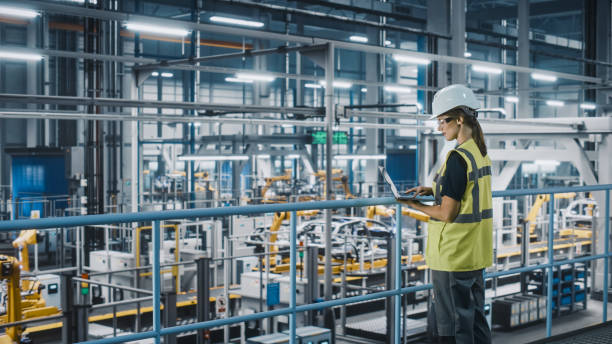 istock Female Car Factory Engineer in High Visibility Vest Using Laptop Computer. Automotive Industrial Manufacturing Facility Working on Vehicle Production with Robotic Arms. Automated Assembly Plant. 1352428671