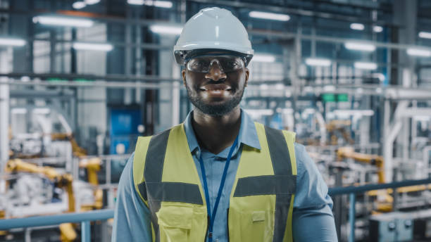 Portrait of an Automotive Industry Engineer in Safety Uniform Smiling at Car Factory Facility. Happy Assembly Plant African American Male Specialist Working on Manufacturing Modern Electric Vehicles. Portrait of an Automotive Industry Engineer in Safety Uniform Smiling at Car Factory Facility. Happy Assembly Plant African American Male Specialist Working on Manufacturing Modern Electric Vehicles. car portrait men expertise stock pictures, royalty-free photos & images