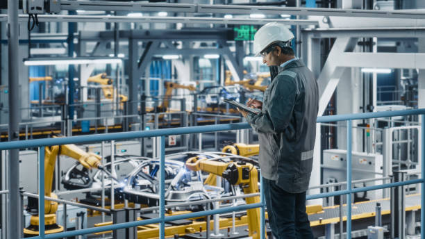 istock Multiethnic Car Factory Engineer in Work Uniform Using Tablet Computer. Automotive Industrial Manufacturing Facility Working on Vehicle Production with Robotic Arms. Automated Assembly Plant. 1352428682