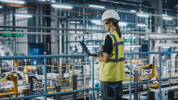 istock Female Car Factory Engineer in High Visibility Vest Using Laptop Computer. Automotive Industrial Manufacturing Facility Working on Vehicle Production with Robotic Arms. Automated Assembly Plant. 1352428701