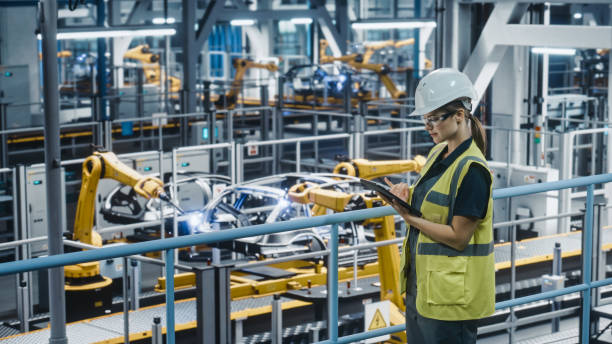 istock Female Car Factory Engineer in High Visibility Vest Using Tablet Computer. Automotive Industrial Manufacturing Facility Working on Vehicle Production with Robotic Technology. Automated Assembly Plant. 1352428711