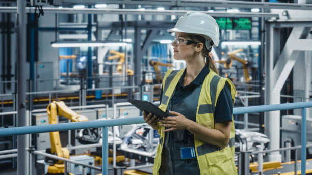 istock Female Car Factory Engineer in High Visibility Vest Using Tablet Computer. Automotive Industrial Manufacturing Facility Working on Vehicle Production with Robotic Technology. Automated Assembly Plant. 1352428715