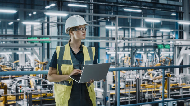 portrait d’une ingénieure de l’industrie automobile 4.0 en uniforme de sécurité utilisant un ordinateur portable dans une usine automobile. spécialiste confiant de l’usine d’assemblage travaillant sur la fabrication de véhicules électriques m - bras robotisé outil de production photos et images de collection