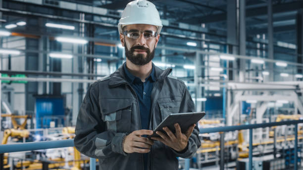 Portrait of Automotive Industry Engineer in Safety Glasses and Uniform Using Laptop at Car Factory Facility. Professional Assembly Plant Specialist Working on Manufacturing Modern Electric Vehicles. retrato del ingeniero de la industria automotriz en gafas de seguridad y uniforme usando computadora portátil en las instalaciones de la fábrica de automóviles. especialista profesional en plantas de ensamblaje que trabaja en la fabricación de vehícul
