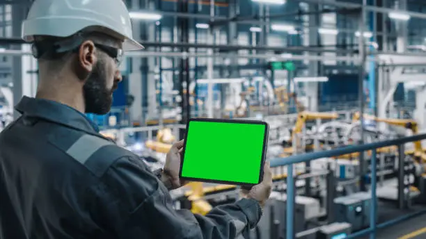 Car Factory Engineer in Work Uniform Using Tablet Computer with Green Screen Mockup Display. Augmented Reality Software at Automotive Industrial Manufacturing Facility Working on Vehicle Production.